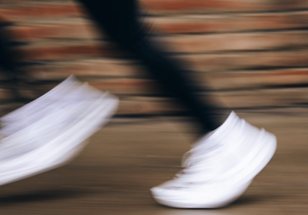 Blurred image of a person running, wearing white sneakers, with a brick wall in the background.